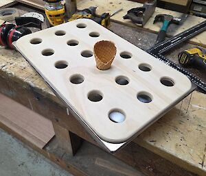 A rectangular wooden board with qith 18 holes in it, one of which holds an ice cream cone