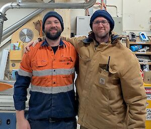 Two men, weatring blue beanies and workshop clothes pose proudly together