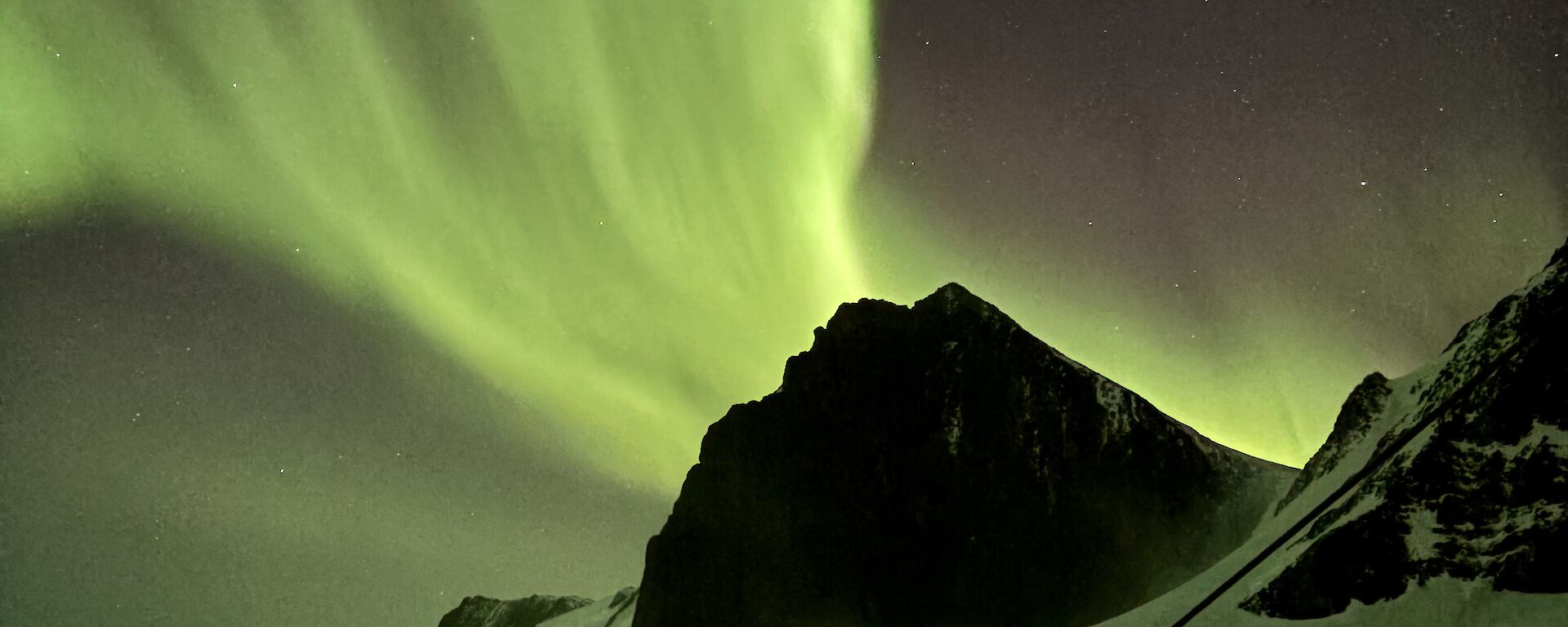 A picture of a bright green aurora overhead, with mountain peaks and sea ice in teh background
