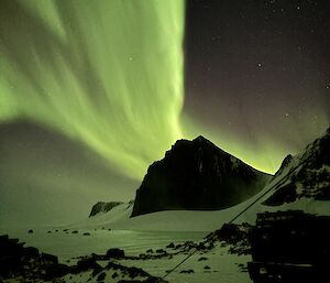 A picture of a bright green aurora overhead, with mountain peaks and sea ice in teh background
