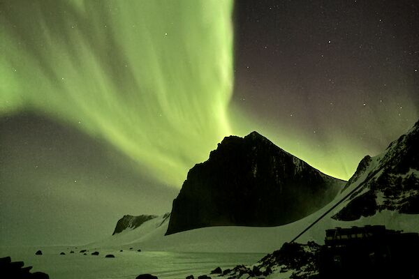 A picture of a bright green aurora overhead, with mountain peaks and sea ice in teh background