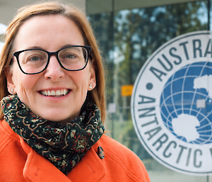 A smiling woman wearing orange with the Australian Antarctic Program logo to her right