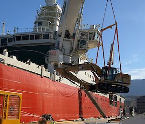 A large excavator being lifted by crane onto a big red ship