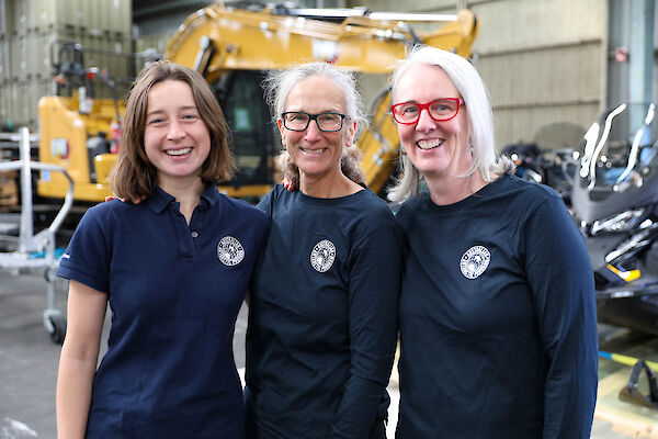 Three women in dark blue Australian Antarctic Program shirts, standing in front of heavy machinery