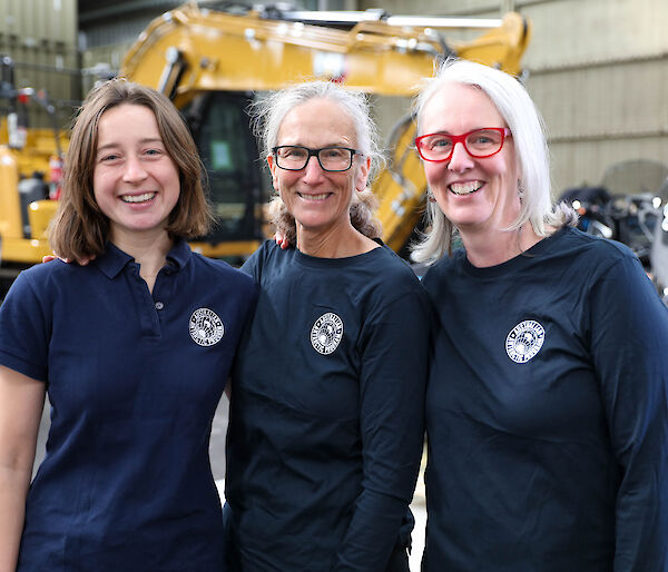 Three women in dark blue Australian Antarctic Program shirts, standing in front of heavy machinery