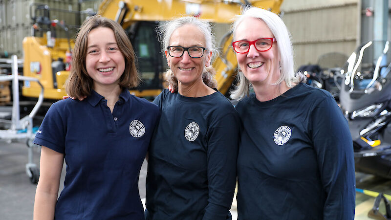 Three women in dark blue Australian Antarctic Program shirts, standing in front of heavy machinery