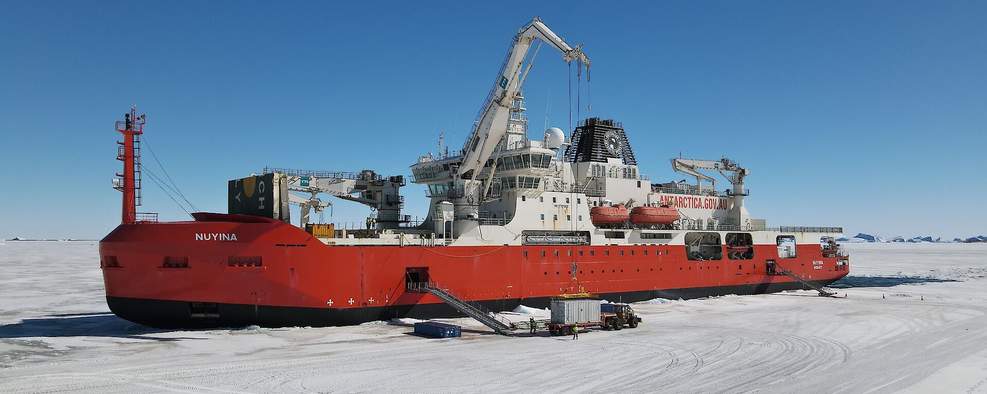 A large red and white ship surrounded by ice with a crane and trucks unloading