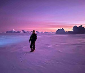 A silhouette of an expeditioner in a snowy landscape coloured with a purple hue, whilst an orange sunset appears behind icebers