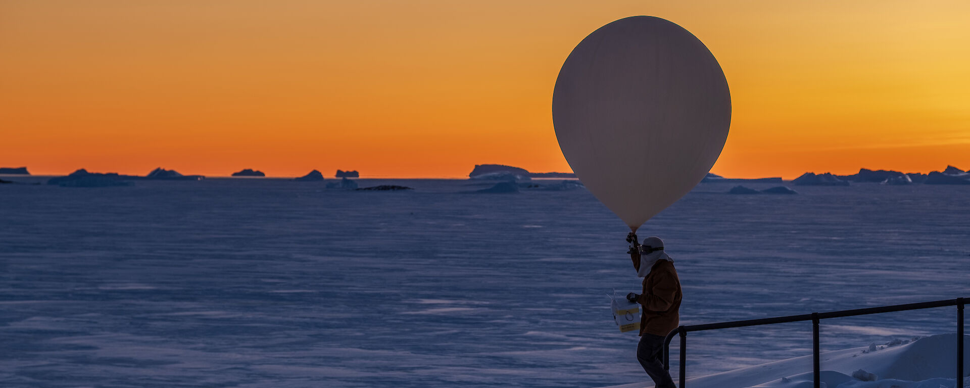 A man holding a large balloon is seen in solhouette in front of and orange sky, with icebergs visible in the distance