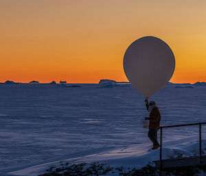 A man holding a large balloon is seen in solhouette in front of and orange sky, with icebergs visible in the distance