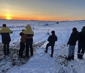 A group of people look towards the horizon over a snowy landscape where the sun is just rising