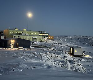 A bright moon hangs in the sky above a series of colourful buildings set in a nsowy landscape