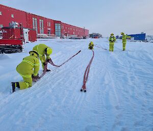 expeditioners wearing firefighting PPE are kneeling and standing in the snow stretching out red fire hoses.