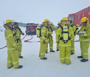 A group of expeditioners in yellow firefighting PPE stand outside of a red building against the snow.
