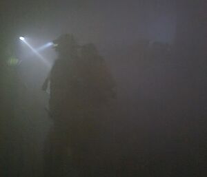 expeditioners in a smoke-filled building. A head torch shines through the smoke.