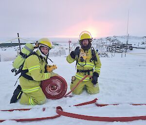 two expeditioners wearing firefighting PPE are kneeling in the snow rolling up red fire hoses.