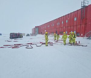 A group of expeditioners in yellow firefighting PPE stand outside of a red building against the snow.
