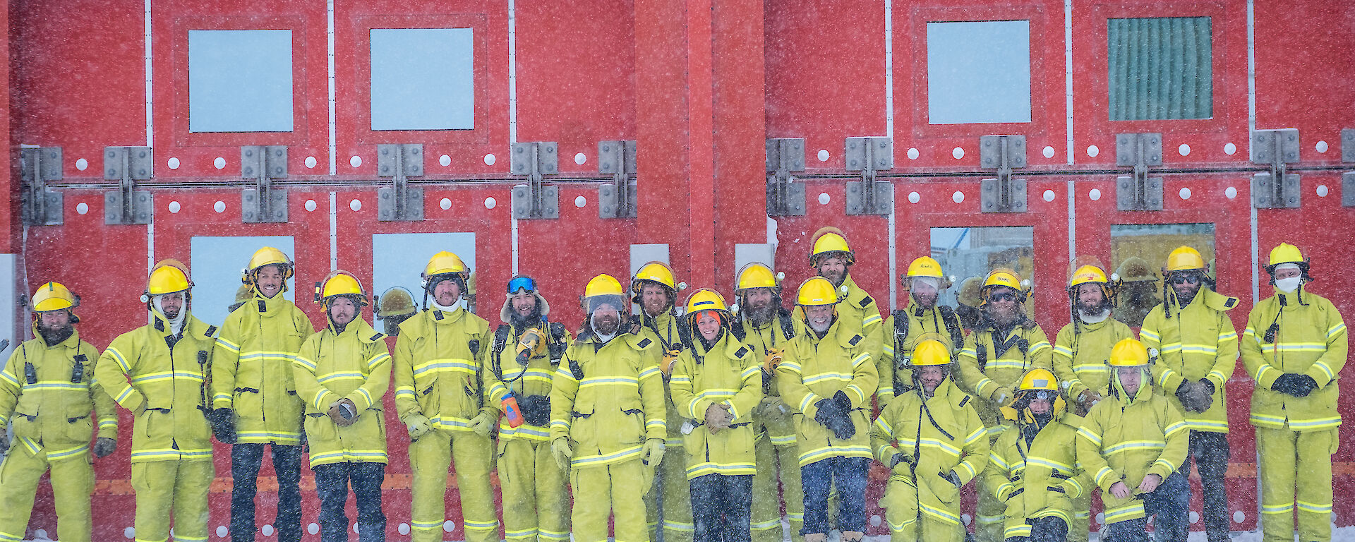 A large group of expeditioners all wearing yellow firefighting PPE, stand in front of a read building.