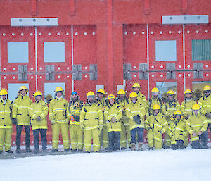 A large group of expeditioners all wearing yellow firefighting PPE, stand in front of a read building.