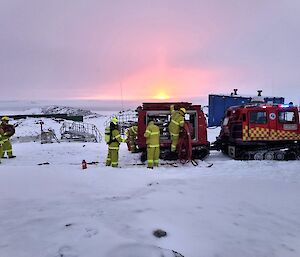 The sun sets in the distance with a pink glow. In the foreground is a red firefighting vehicle and firefighter in yellow PPE