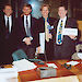 Four people in suits stand behind a desk.