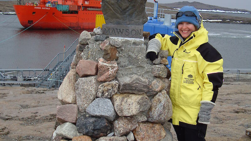 Female expeditioner posing next to statue of Mawson at the station, with icebreaker in the background