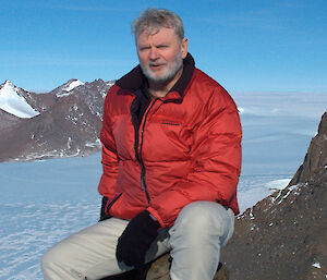 Davis Station Leader, Graham Cook sits on a rocky slope in Antarctica and faces the camera