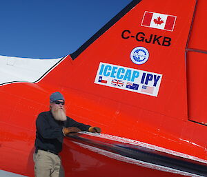 Bearded expeditioner wearing sunglasses and beanie, leans against Basler aircraft