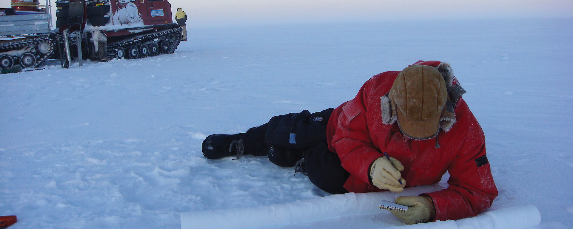 Ice core chemist lying on the ice, cataloguing ice cores, with vehicle in background