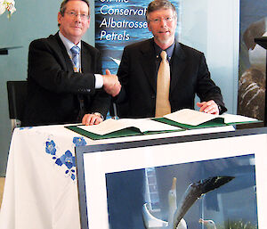 Two men shaking hands whilst signing an agreement