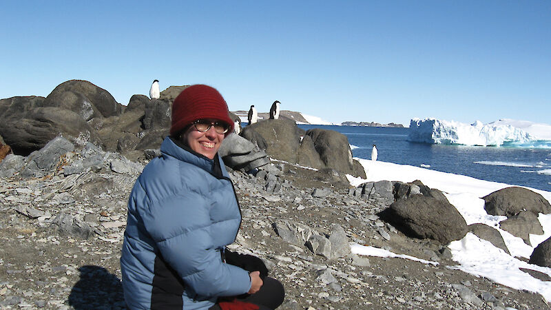 Lynette Finch seated on rocky ground with penguins and icebergs in the background