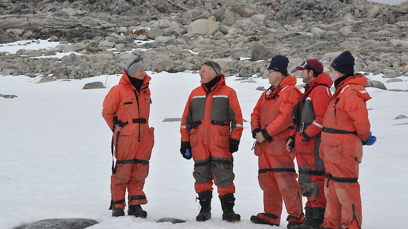 Dr Tony Press and three others, standing in orange Antarctic freezer suits, receive a briefing from a scientist at Casey station