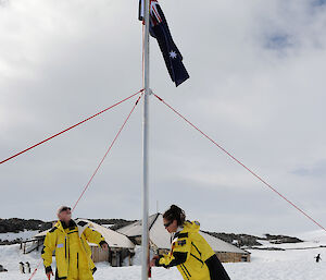 Former expeditioner and president of ANARE, David Ellyard (left), and Deb Bourke (right), raise the Australian flag in front of Mawson’s Huts, in celebration of the 100th anniversary of Mawson’s Landing at Commonwealth Bay in Antarctica.