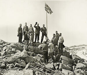 Mawson (with hat raised) and his group of expeditioners standing on rocks with British flag erected