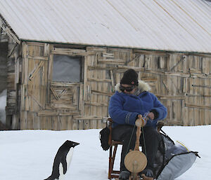 Marion Wheatland is inspected by a curious Adelie penguin while spinning wool in front of Mawson’s Huts.