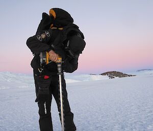 An expeditioner using an electric drill and auger to drill into the sea ice — pale, pink sky and mountains in the background.