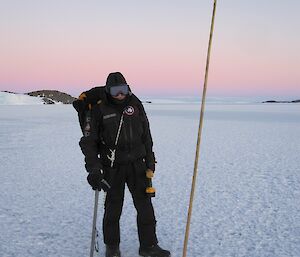 An expeditioner standing beside a cane on the sea ice. There is a pale, pink sky and mountains in the background.