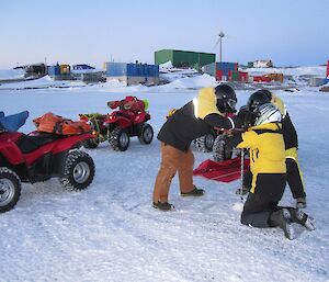 Three people drilling through the sea ice with a hand auger. There are three quad bikes beside them parked on the sea ice.