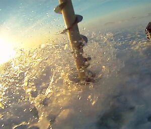 A close up of a stainless steel auger about a metre long vertically drilling into sea ice with a spray of sea water coming up out of the hole