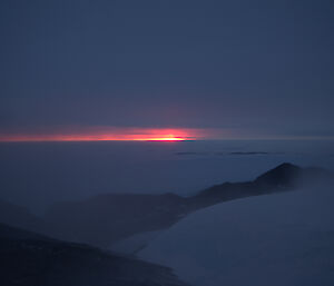 In the foreground are the rocks and ice with drifting snow and in the background the frozen ocean and a setting sun