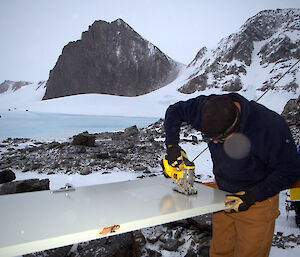 The carpenter outside the hut cutting a section from the door in order to insert the window with ice and mountains in the background