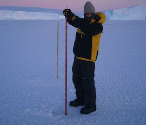An expeditioner about to lower his measuring gauge into a drilled hole in the sea-ice, the ice cliffs of West Bay are in the background