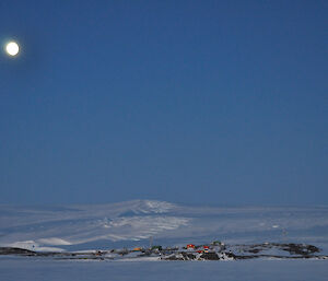 A nearly full moon rising over the buildings of Mawson in the foreground with the icy Antarctic plateau behind