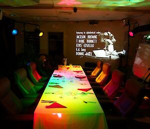 A table set up for 7 diners with a black and white projected on the screen and coloured lights on the ceiling