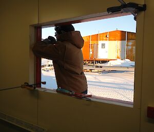 An expeditioner fitting a window in a new building with snow, site service pipes and another building behind