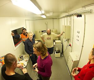 Six expeditioners in the Detector Room. One has his left hand on the small decay cabinet on the wall and is standing in front of the 2 detectors