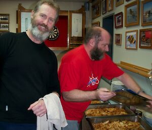 Two expeditioners standing beside several trays of fish and chips prepared for dinner