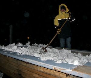 An expeditioner in winter clothes holding a shovel with the outside hot tub full of snow in the evening dark