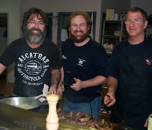 Three expeditioners standing behind the griller on which onions and sausages are cooking