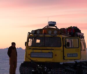 An over-snow vehicle with person beside it and pastel colours of sunrise